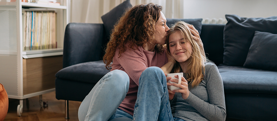 White mom kisses head of daughter while sitting on floor with coffee mug