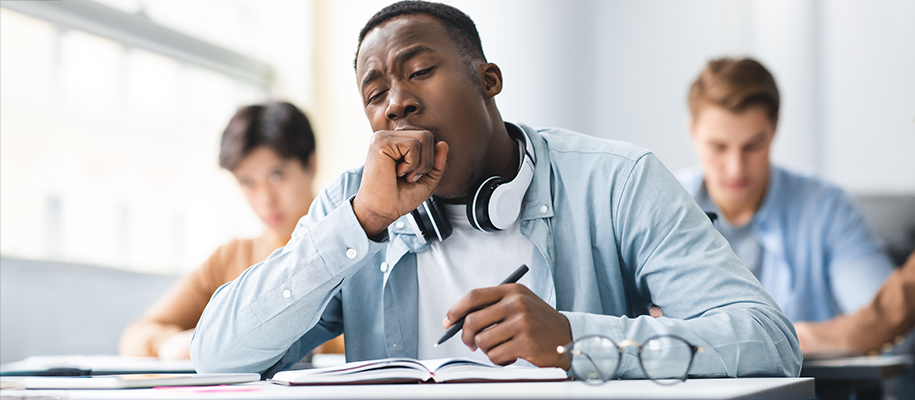 Black male student with white headphones around neck yawning at desk in class