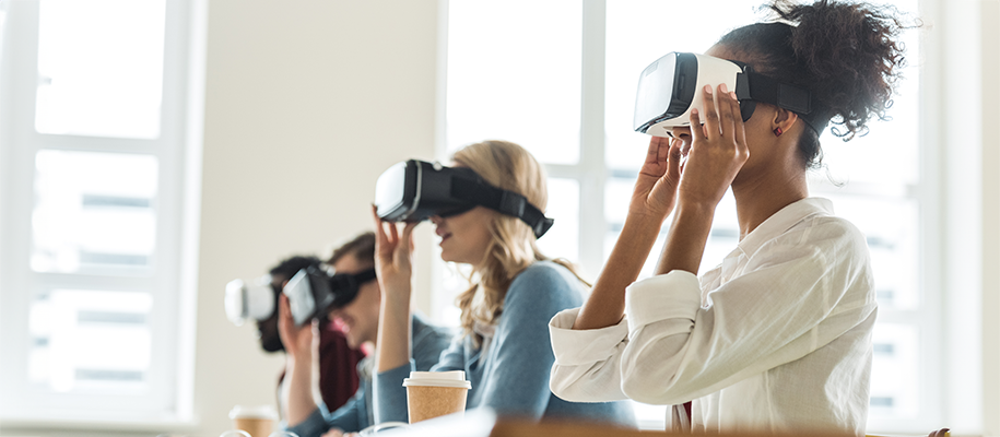 Four diverse students wearing VR goggles in desk row of college class