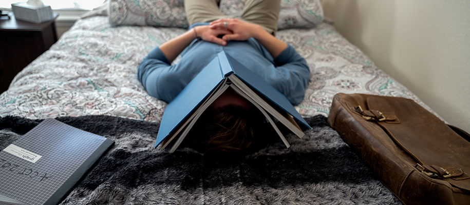Man in blue shirt asleep under open book on bed with back and notebook