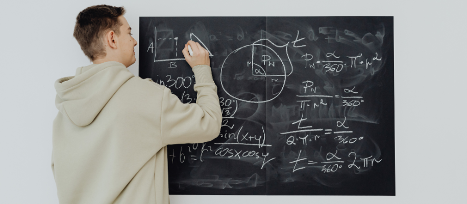 White male teen in beige hoodie doing math equations on small chalkboard