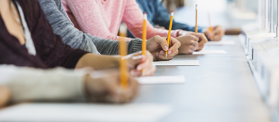 Focus on desk where line of students hands hold pencils over exam sheets