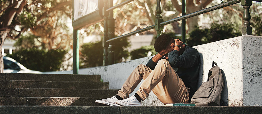 Black student sitting with head down, hands on neck, next to backpack on campus