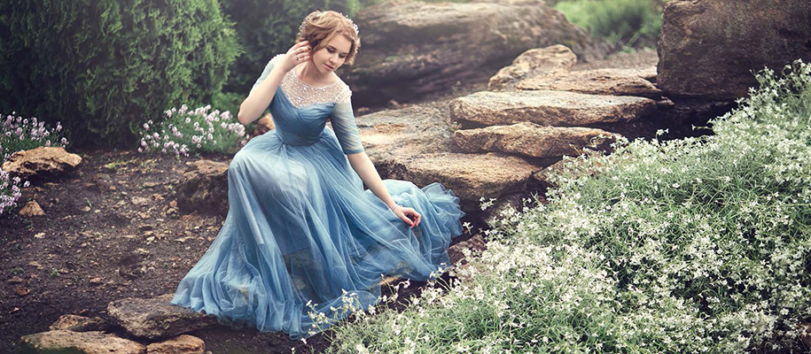 Girl in blue dress and done up hair crouching on stone path near flowers