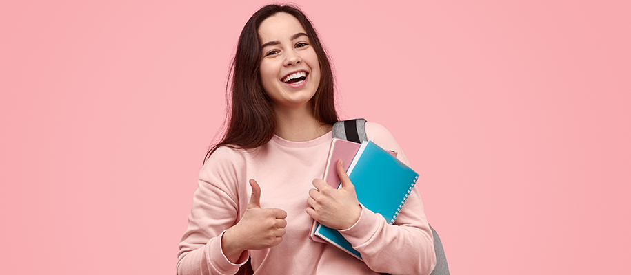Young woman in pink sweatshirt carrying books, smiling and giving thumbs up