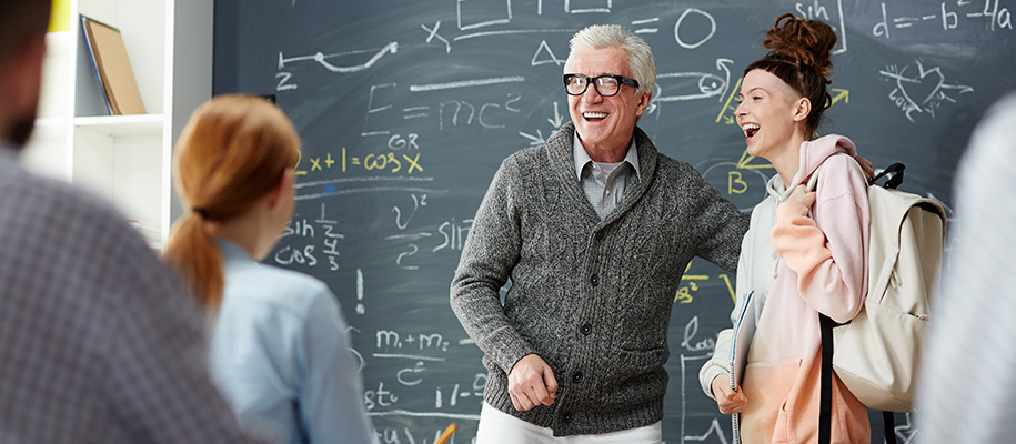 Older White man in sweater laughs with student in sweatshirt in front of class