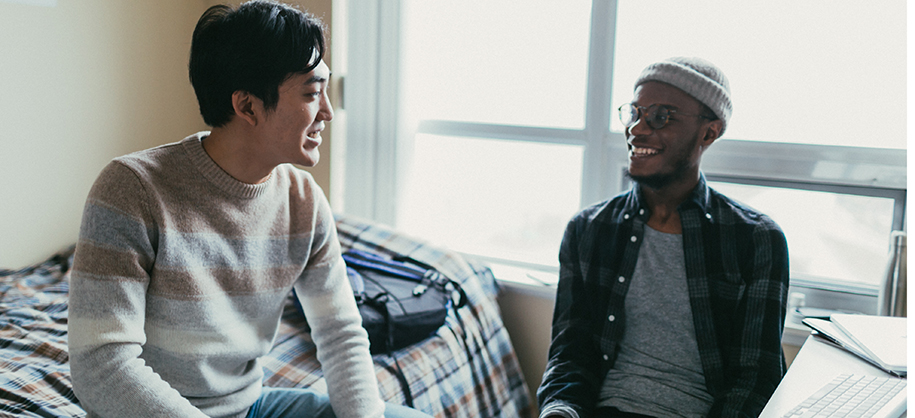 Young Asian man in sweater and Black man in dorm room smiling at each other