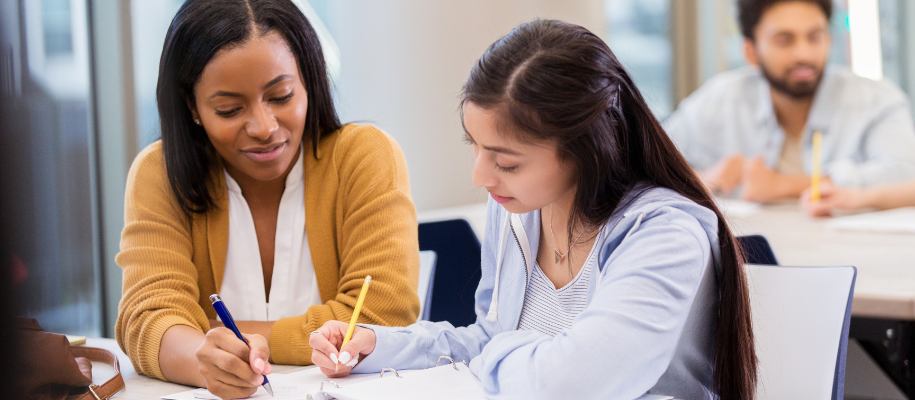 Black woman in sweater helping young Hispanic woman with assignment in class