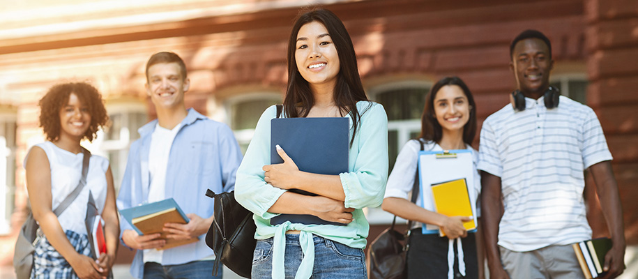 Five diverse students with books and bags outside, focus on Asian woman in front