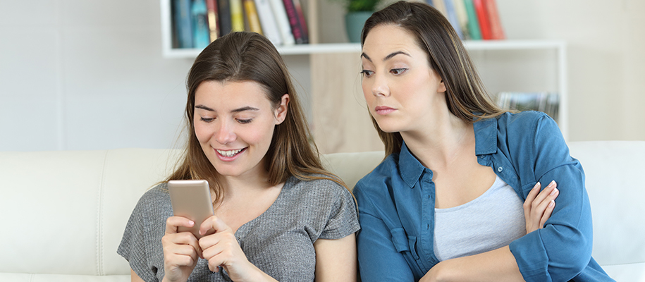 Young White woman on couch, White roommate leaning over spying on phone