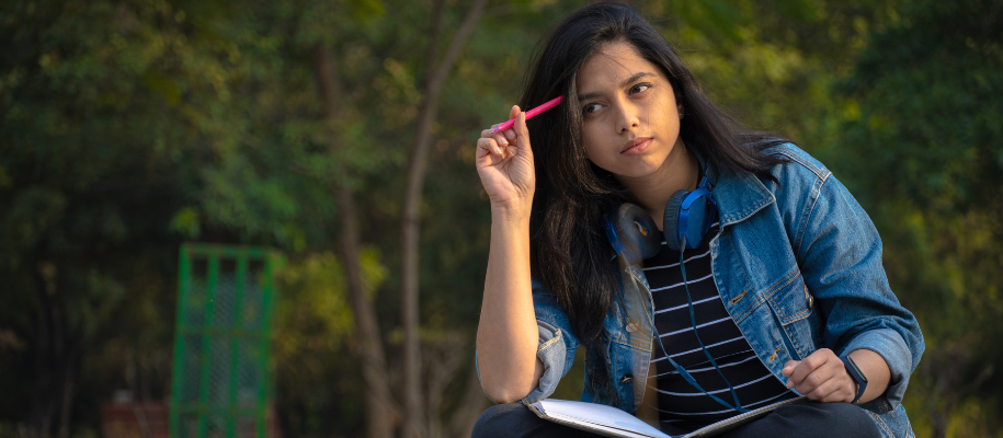 Indian woman in denim shirt sitting in grass with notebook and pink pen to head