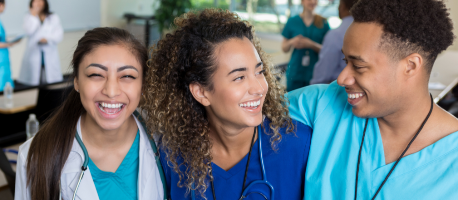 Three diverse medical students in various colored scrubs, smiling in classroom