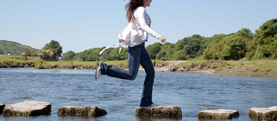 Young woman in white sweater and jeans jumping across rocks on water