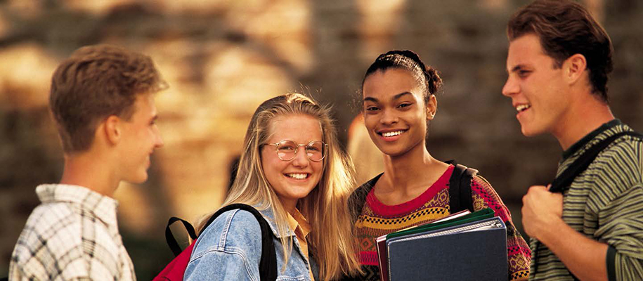 Two White men, White woman, Black woman in front of brick wall with backpacks