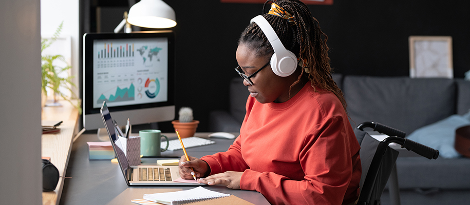Black woman in a wheelchair with white headphones doing business work at desk