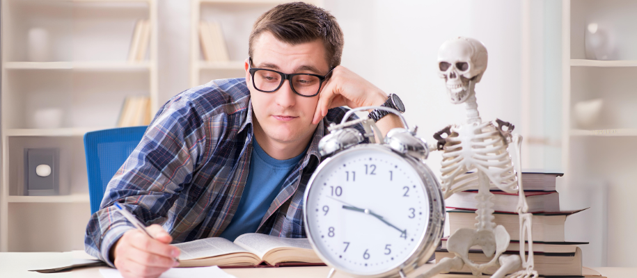 White man in plaid shirt, glasses studying, skeleton model sits by alarm clock