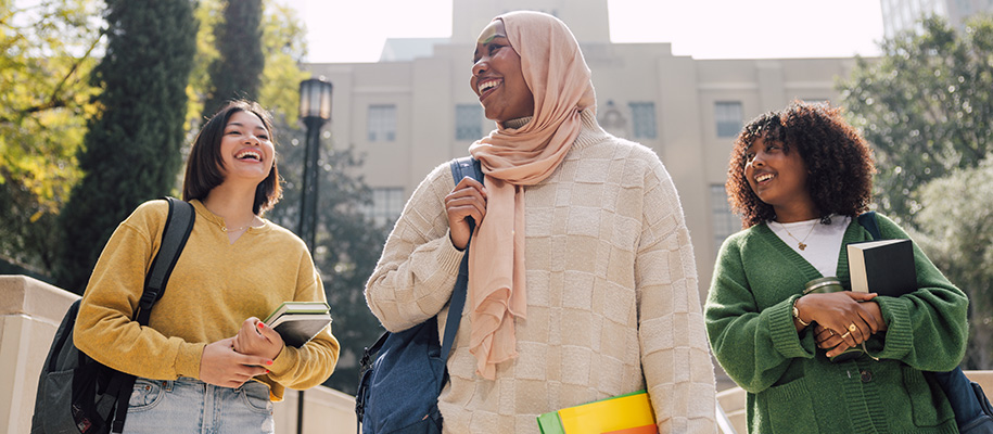 Asian woman and two Black women, one in a hijab, in sweaters on college campus