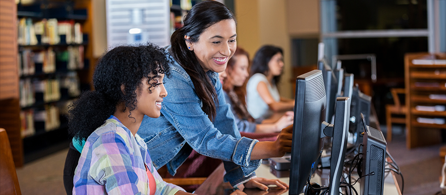 Hispanic counselor helping Black teen girl in plaid on computer in library