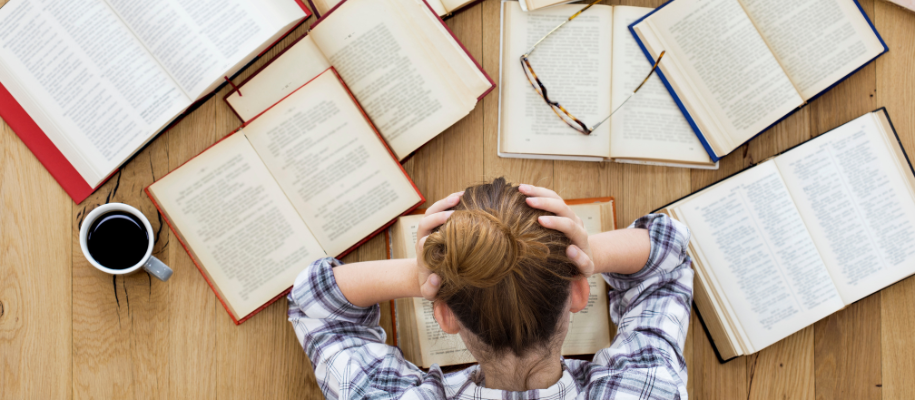 Overhead of woman in blue plaid shirt holding head, surrounded by books, coffee