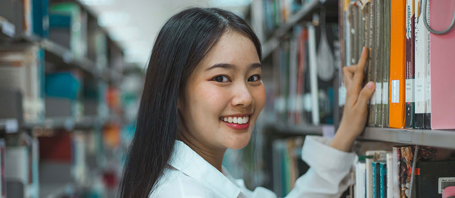 Asian woman with long black hair smiling at camera while perusing library shelf