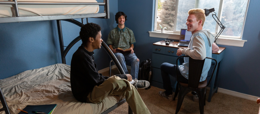 Three male roommates sitting in desk chairs, on bed in dorm room with blue walls