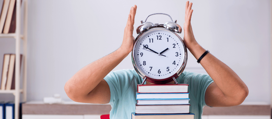 Person behind a stack of books holding an alarm clock in front of their face