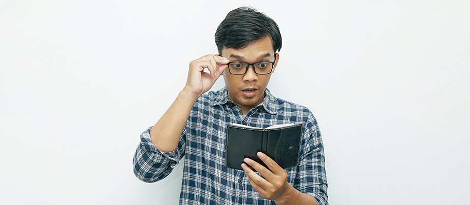 Indonesian man looking at black checkbook and holding glasses on face in shock