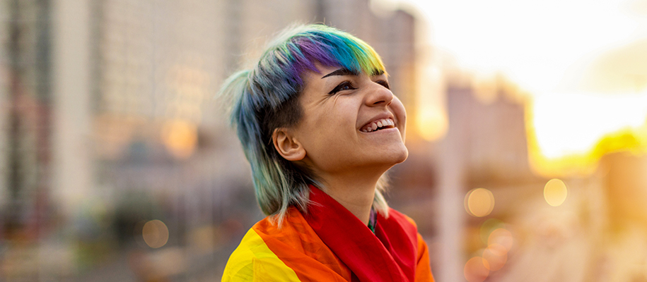 Gender fluid person with rainbow hair, flag around shoulders, smiling at sunset