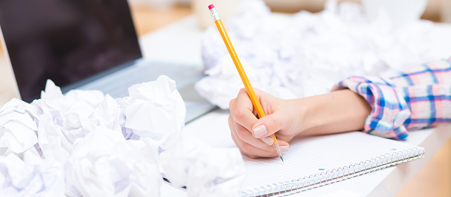 Arm of White person in plaid shirt holding pencil over notebook surrounded by papers crumpled into balls