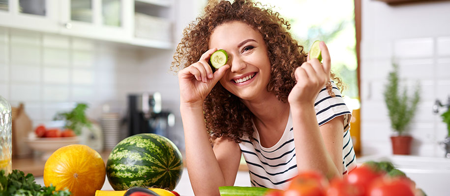 Curly-haired female smiling in kitchen with veggies and cucumber slice on eye