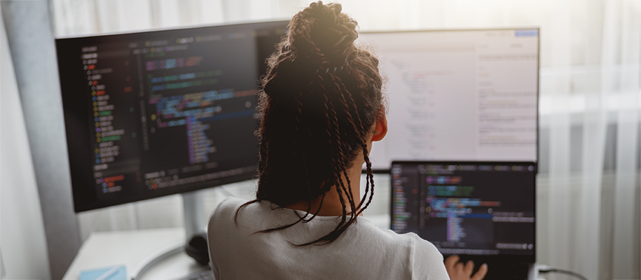 Black woman with bun of braids in front of three screens of coding software
