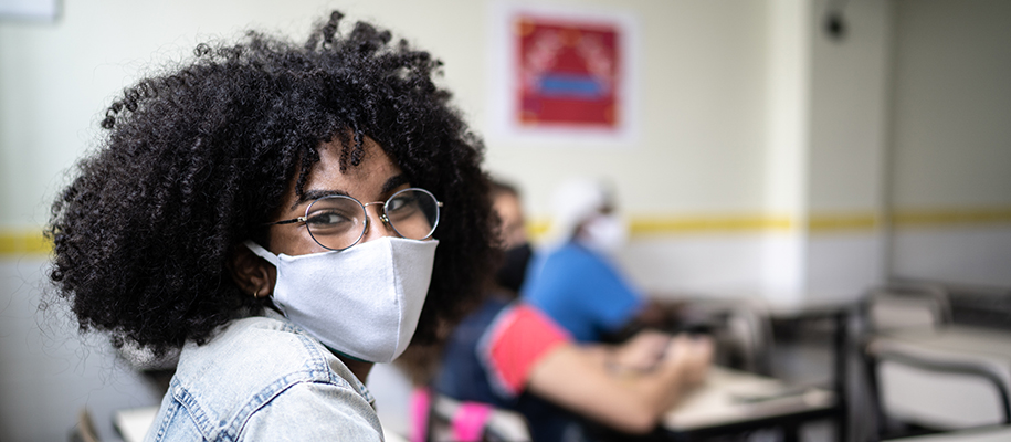 Young Black woman with Afro, circular glasses, white face mask, in classroom