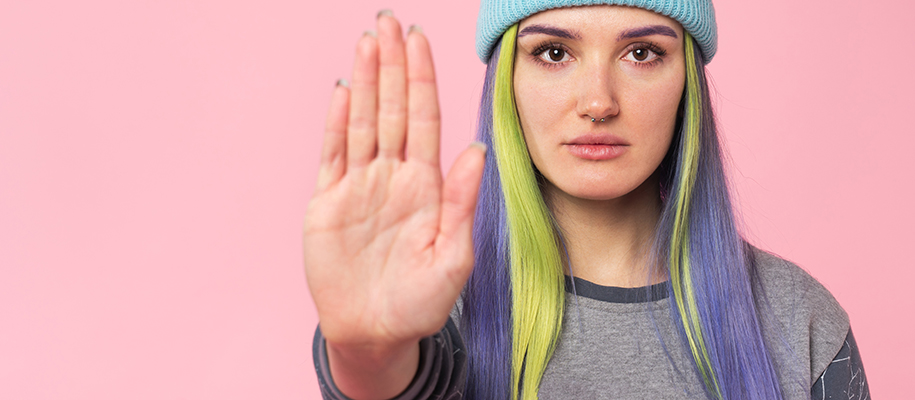 White woman with colorful hair, nose piercing, hat, holding palm up in halt sign