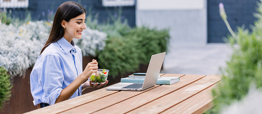 White woman in blue button-up shirt eats salad on laptop outside at picnic table