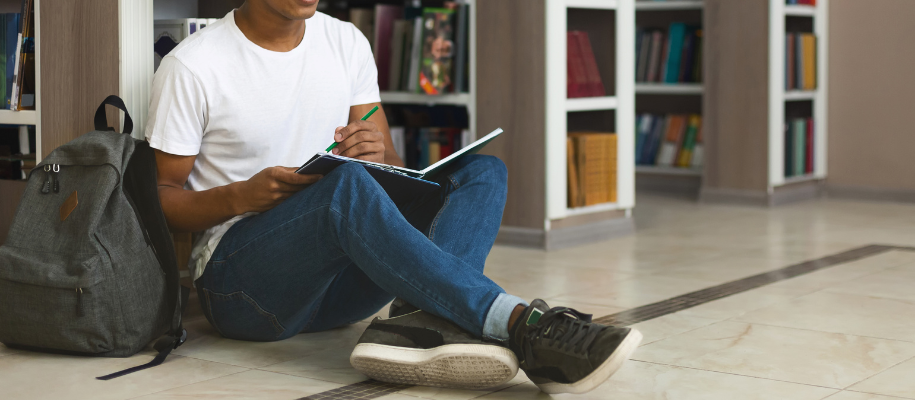 Black student in T-shirt, jeans sitting on library floor, writing in notebook