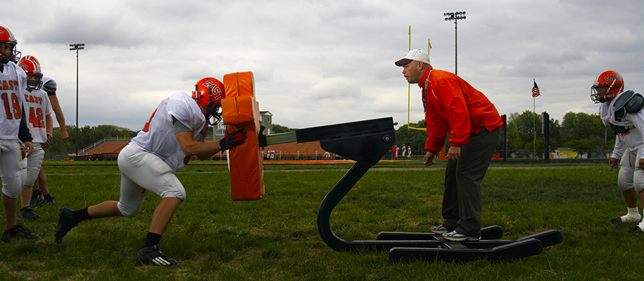 Teen football player in white and orange pushing block sled with coach on it