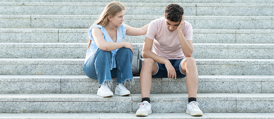 White woman in blue shirt, jeans comforting White man looking sad on steps