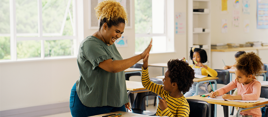 Young Black woman teacher in green shirt high-fives a young Black boy in yellow