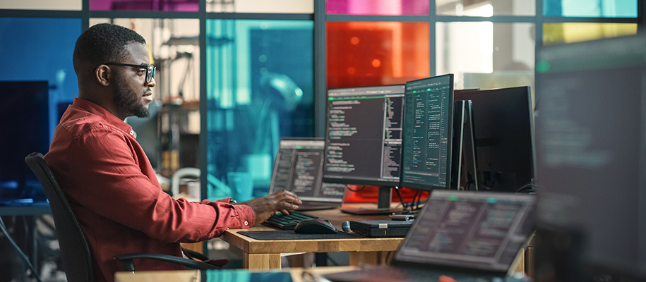 Black man in red shirt coding on multiple monitors in front of colored glass