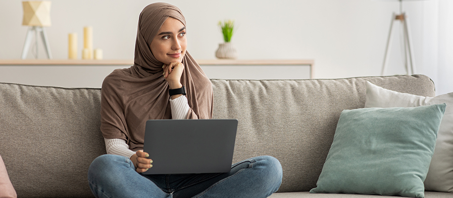 Woman in brown Al-Amira sitting on couch with laptop, head on fist, thinking