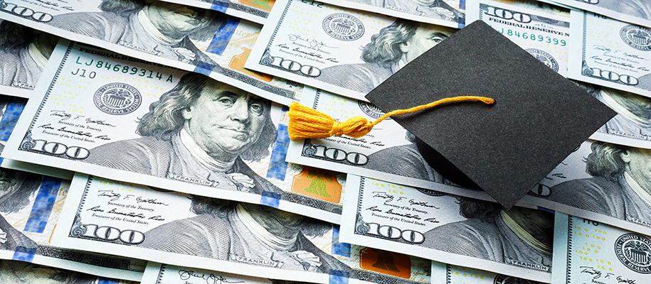 Overlapping layer of hundred dollar bills with black grad grad cap on top
