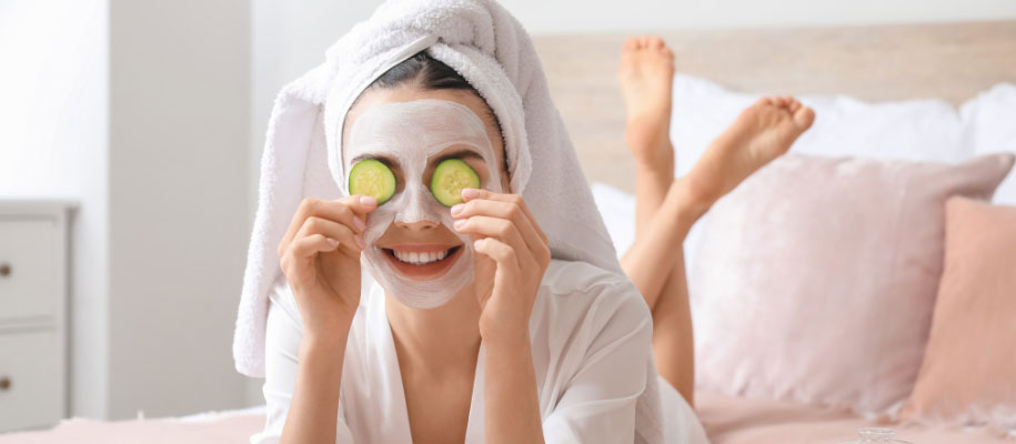 White woman lying on bed with legs crossed, hair in towel, cucumbers on eyes