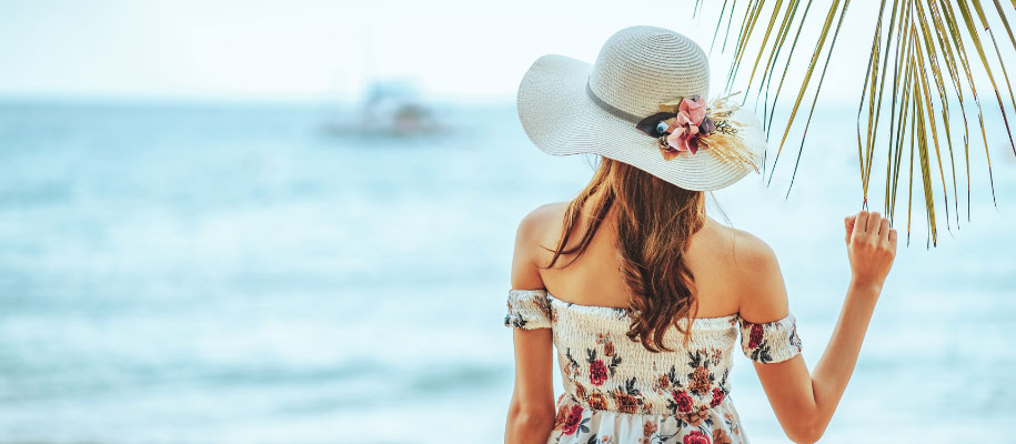 Back of woman in sundress and sunhat, touching palm tree, looking at ocean