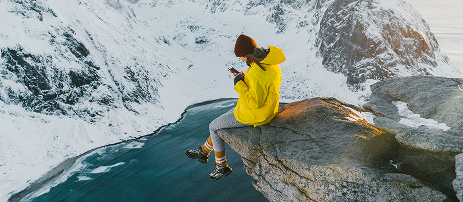 Woman in yellow winter coat and hat, texting on cliff edge in snowy mountains