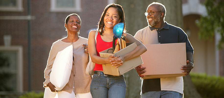 Black student in red shirt carrying box, parents carrying pillow and box behind