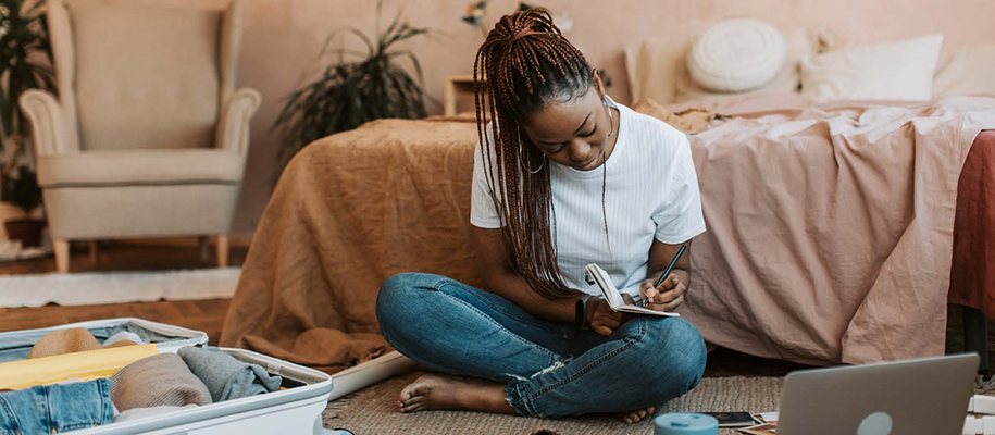Black woman with braids, sitting on floor packing and writing in notebook