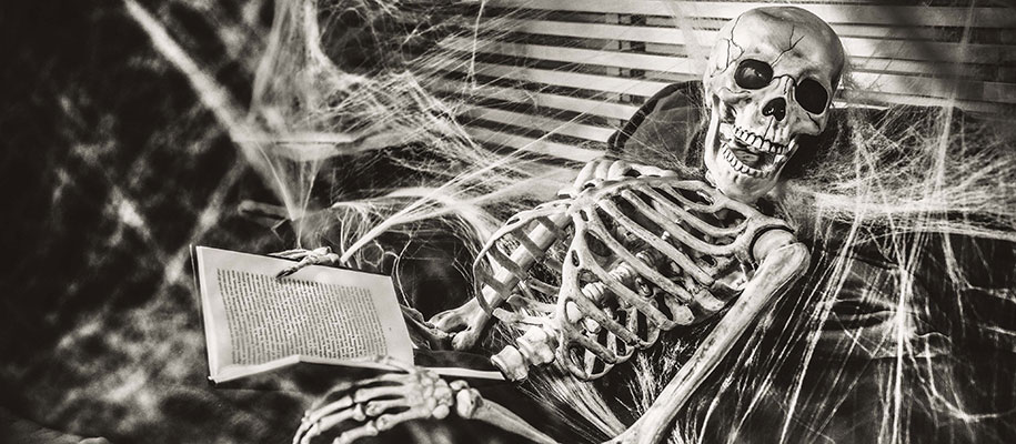 Black and white shot of skeleton holding book sitting in chair with cobwebs