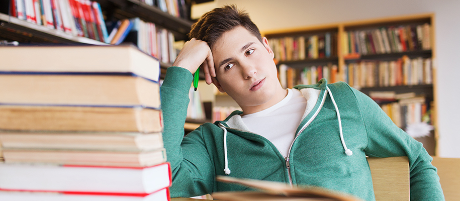 White male in green sweatshirt in library looking at stack of books bored