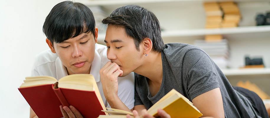 Two Asian men lying on bed together reading books, shelf with books behind