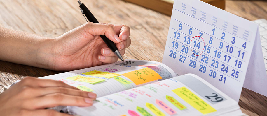 Hands writing in planner covered in colored tabs, next to paper calendar